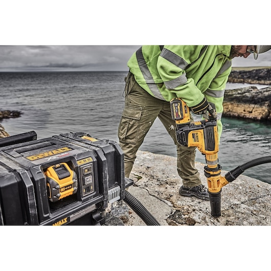 A worker in a green safety jacket uses a yellow DEWALT power tool to drill into concrete next to a body of water, with a