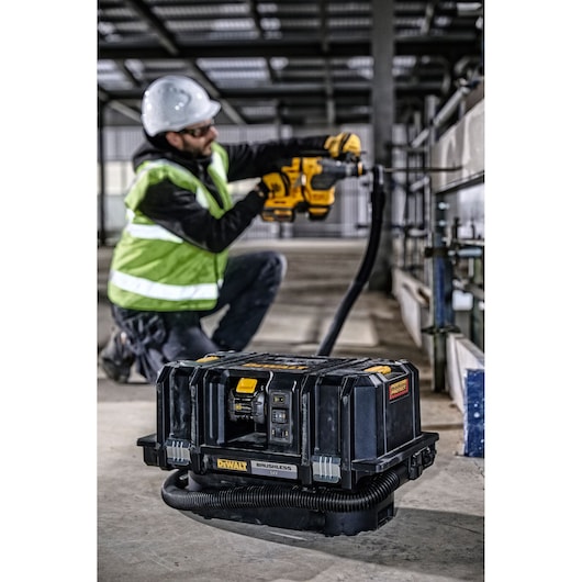 A DEWALT DCV586MT2 brushless vacuum cleaner is placed on a construction site floor, with a worker in a safety vest and