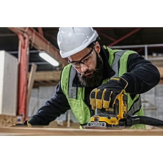 A person wearing a hard hat, high-visibility vest, and protective gloves is using a DEWALT DCW200P2 cordless sander on a