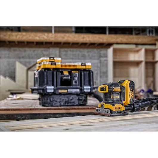 A DEWALT cordless sander (DCW200P2) is placed on a wooden surface, with a DEWALT tool storage box in the background. The