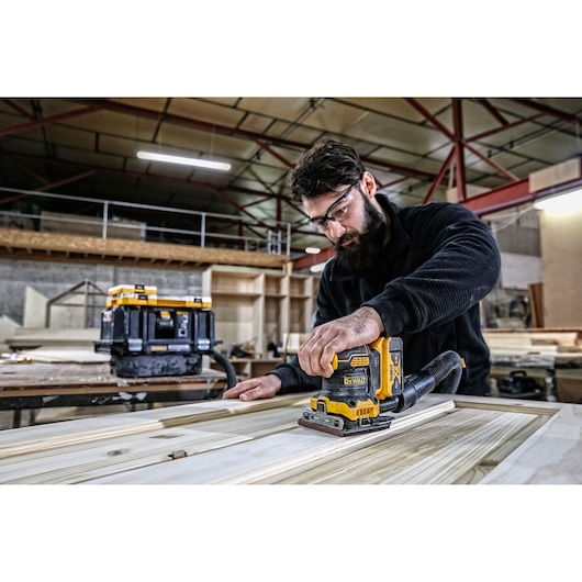 A person using a DEWALT DCW200P2 electric sander to smooth a wooden surface in a workshop. The person’s face is blurred, and