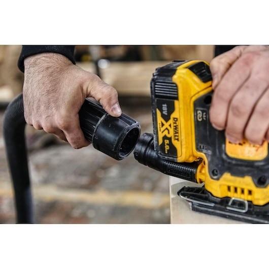 Hands connect a vacuum hose to a yellow DEWALT tool for dust extraction in a workshop setting.