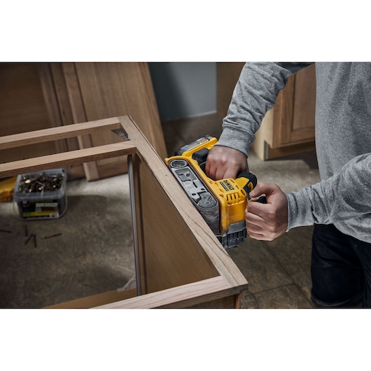 A person uses a yellow DEWALT DCW220N cordless belt sander to smooth the edge of a wooden frame in a workshop setting.