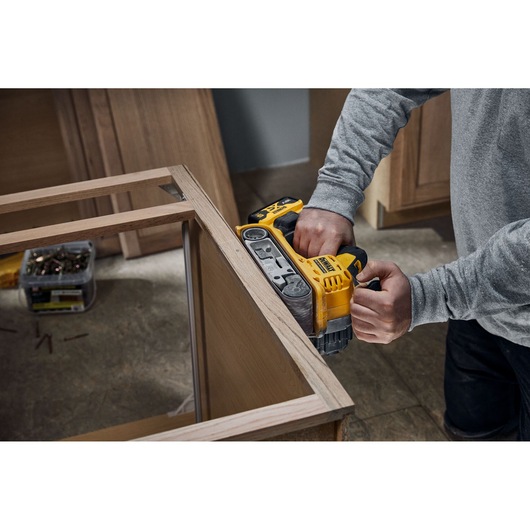A person using a yellow DEWALT cordless belt sander to smooth the edge of a wooden frame in an indoor workspace.