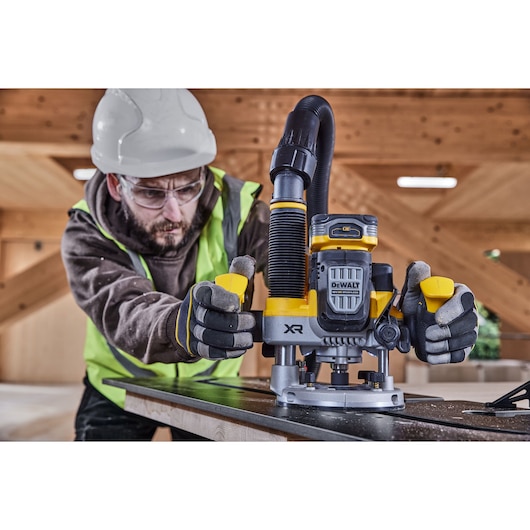 A construction worker wearing a safety helmet, gloves, and a high-visibility vest is operating a DEWALT DCW620H2 cordless plunge router on a piece of wood, with a dust extraction hose attached for clean operation. The workspace appears to be a wooden structure under construction.