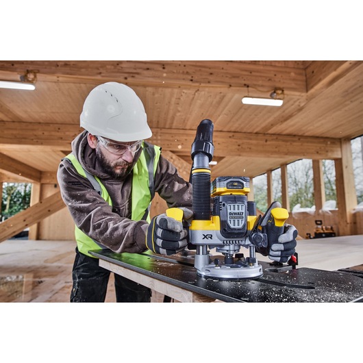 A person wearing safety equipment operates a DEWALT XR cordless router (model DCW620H2) on a black work surface in a wood construction area.