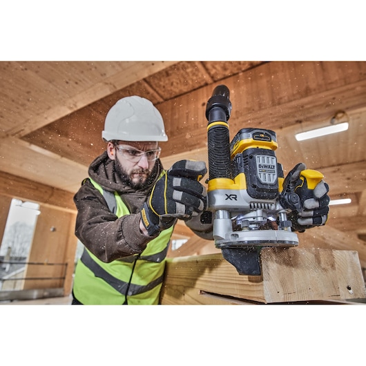 A worker wearing a safety helmet and high-visibility vest uses a DEWALT XR cordless router to work on a piece of wood inside a wooden structure.