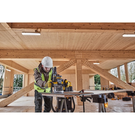 A person wearing a white hard hat and yellow safety vest is operating a DEWALT power tool on a workbench inside a wooden construction building.