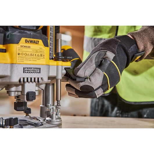 Gloved hand adjusts a DEWALT DCW620 router tool on a wooden surface in a workshop setting.