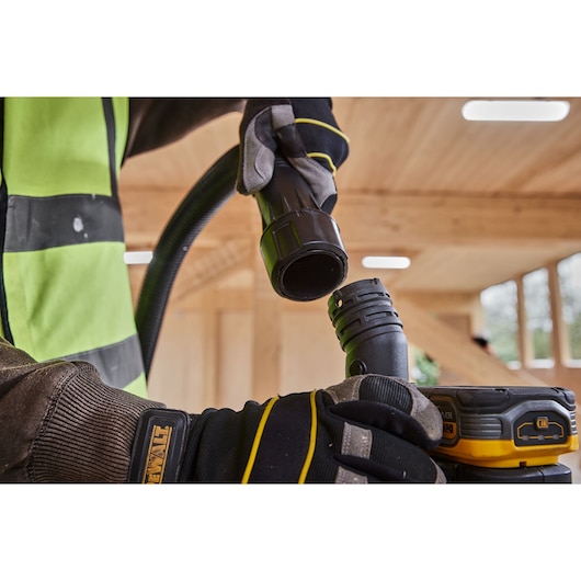 Worker connecting DEWALT vacuum hose to tool in a wooden workspace, wearing gloves and safety vest.