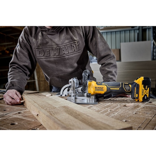 DEWALT DCW682P2-QW, DCW682P2 tool on wood in workshop. Person wears DEWALT sweatshirt. Photography product image.