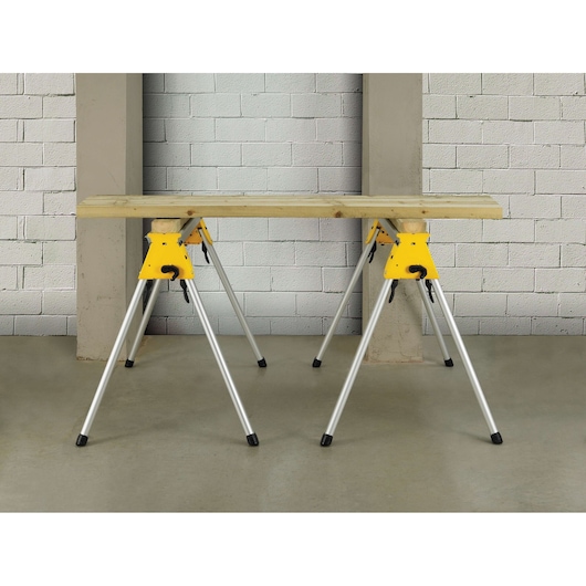 A pair of DEWALT folding metal sawhorses with yellow brackets and silver legs, supporting a wooden plank in an indoor workshop setting.