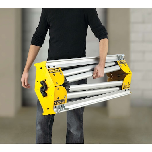A person holding a folded DEWALT DE7035 heavy-duty metal stand with yellow end panels and silver bars, suitable for supporting tools such as miter saws.