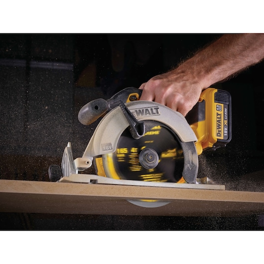 A DEWALT cordless circular saw in use, cutting through a wooden board. A person's hand is holding the tool, and sawdust is