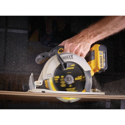 A DEWALT 18V cordless circular saw being used to cut a piece of wood, held by a person's hand. Sawdust is visible as the saw