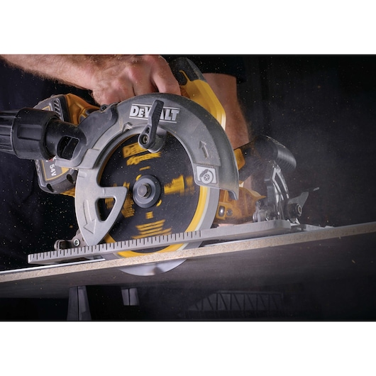 A close-up of a DEWALT circular saw in use, cutting through a piece of wood, operated by a person's hand.