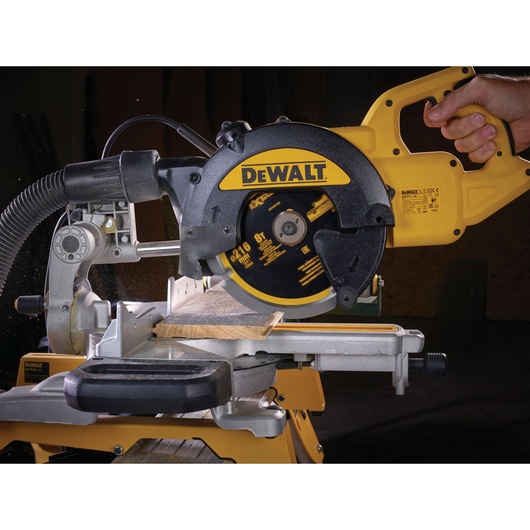 A close-up view of a DEWALT branded electric miter saw cutting a piece of wood, with a hand operating the tool. The saw