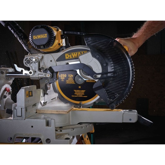 A close-up view of a DEWALT compound miter saw cutting a piece of wood. The saw blade has a protective cover and a person is