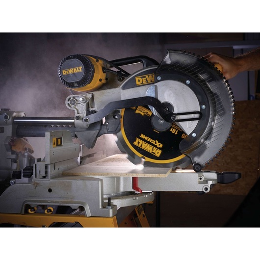 A DEWALT mitre saw cutting a piece of wood, with visible dust and a person's hand operating the tool.