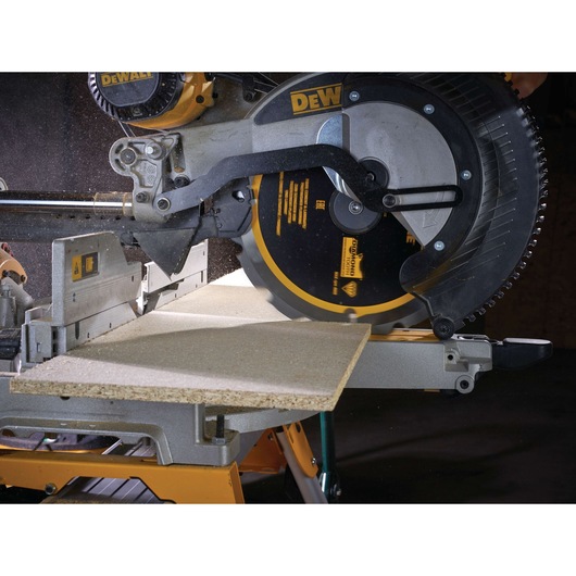 A close-up image of a DEWALT miter saw cutting a piece of particle board. The saw blade is active and surrounded by safety