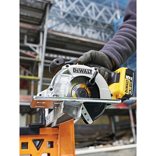 A close-up view of a DEWALT cordless circular saw in use, cutting through a metal piece. The tool is held by a gloved hand
