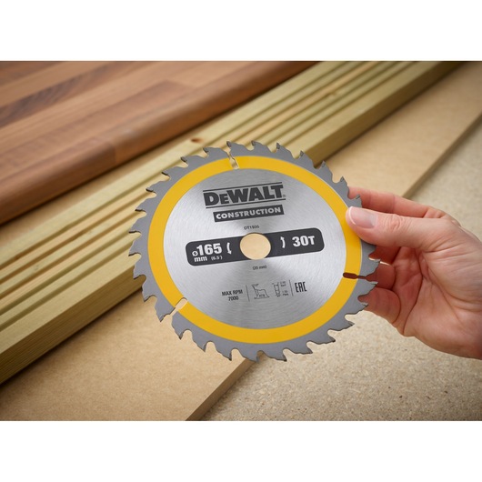 Hand holds DEWALT DT1935 circular saw blade, labeled for construction, in front of wood planks and boards.