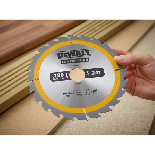 A hand holding a DEWALT Construction circular saw blade, model DT1944, with 24 teeth and a diameter of 190 mm, against a background of wood planks.
