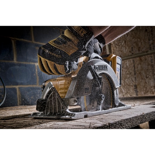 Close-up of a DEWALT cordless circular saw cutting through a piece of wood, operated by a person wearing protective gloves. Sawdust is visible in the air.