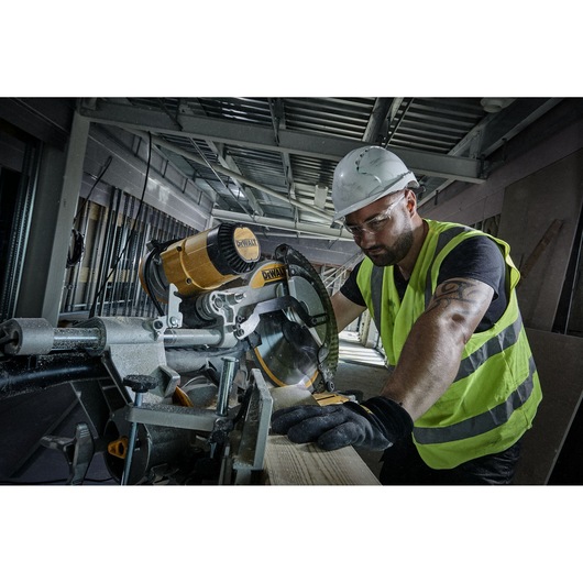 A construction worker wearing a high-visibility vest and helmet is using a DEWALT miter saw to cut a piece of wood inside a