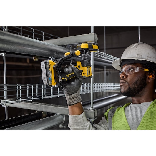 A construction worker in safety gear uses a DEWALT cordless tool to drill into a metal surface on an industrial site. The