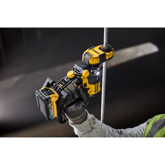 A close-up view of a gloved hand using a DEWALT cordless power tool to cut a metal rod. The tool is yellow and black, with