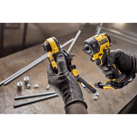 A close-up of gloved hands holding a DEWALT branded impact driver and an attachment accessory. The tools are positioned above a workbench with metal rods and cylindrical parts scattered on the surface.