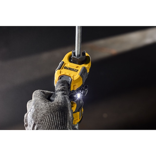 A close-up of a DEWALT power tool being used by a gloved hand to cut a metal threaded rod. The tool is yellow and black, with a built-in LED light illuminating the work area.
