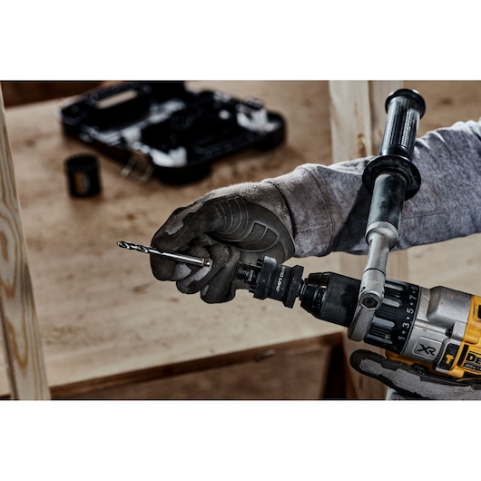 A close-up of a person wearing work gloves inserting a drill bit into a DEWALT power drill, with a blurred background of tools and wood.