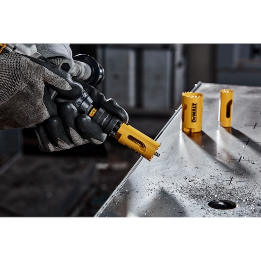 A gloved hand is using a DEWALT hole saw to drill into a piece of metal. Two additional DEWALT hole saw attachments are placed on the metal surface, and metal shavings are visible around the drilled hole.