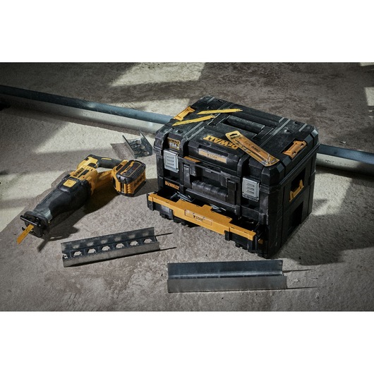 A DEWALT reciprocating saw, battery pack, metal cutting blades, and a black and yellow DEWALT tool storage box placed on a concrete floor. Construction materials and metal pieces are arranged around the tools.