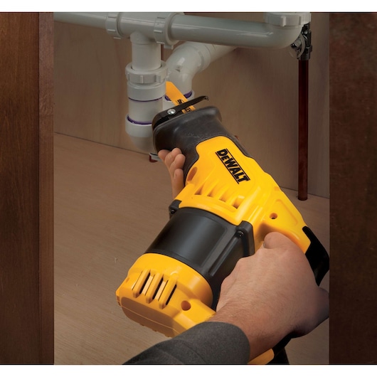 A DEWALT reciprocating saw being used to cut a plastic pipe beneath a sink, with hands holding the tool.