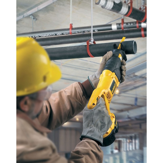 A worker wearing safety gear uses a DEWALT reciprocating saw to cut through a black pipe suspended from the ceiling in an