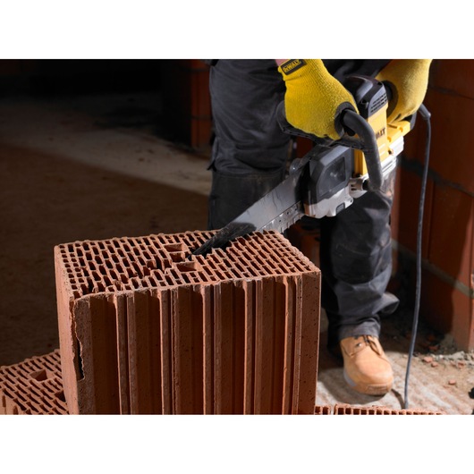A person wearing yellow gloves and work boots is using a DEWALT electric saw to cut through a large, hollow construction brick.