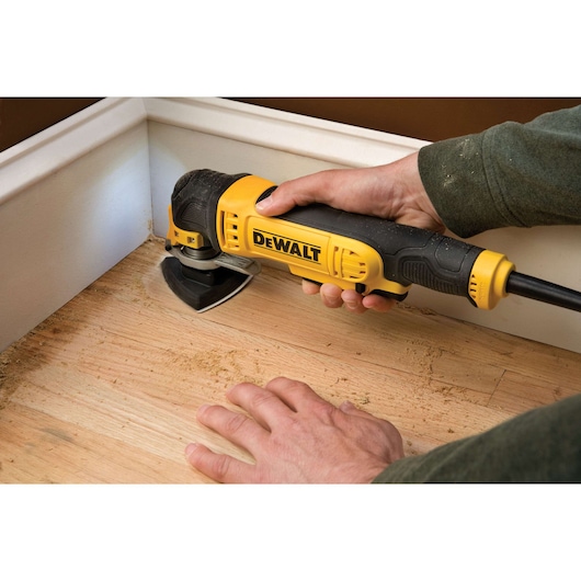A person is using a DEWALT oscillating multi-tool to sand or work on a wooden floor near a wall and baseboard.