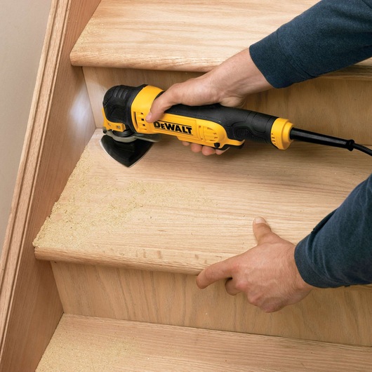 Person sanding wooden stairs with a DEWALT electric tool, dust visible on the steps.