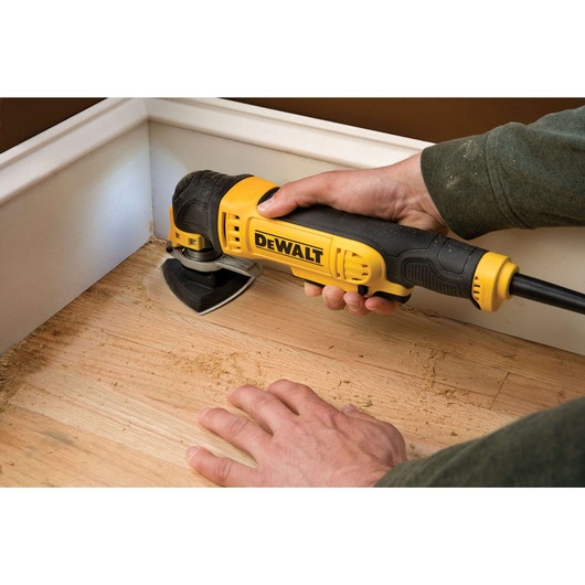 A person uses a yellow and black DEWALT multi-tool to sand the wooden floor near a wall corner.