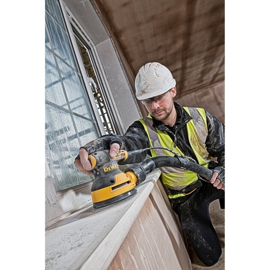 A construction worker wearing a safety vest and helmet uses a DEWALT DT3103 power sander on a window ledge inside a building
