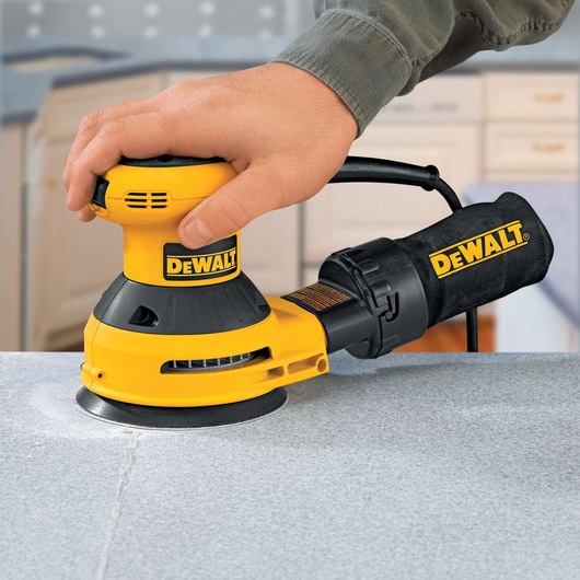 A DEWALT DT3115 orbital sander being used to sand a flat surface, with a person's hand holding the tool. The sander is