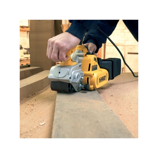 A person is using a DEWALT belt sander to smooth a wooden plank on a work surface, with wood shavings visible around.