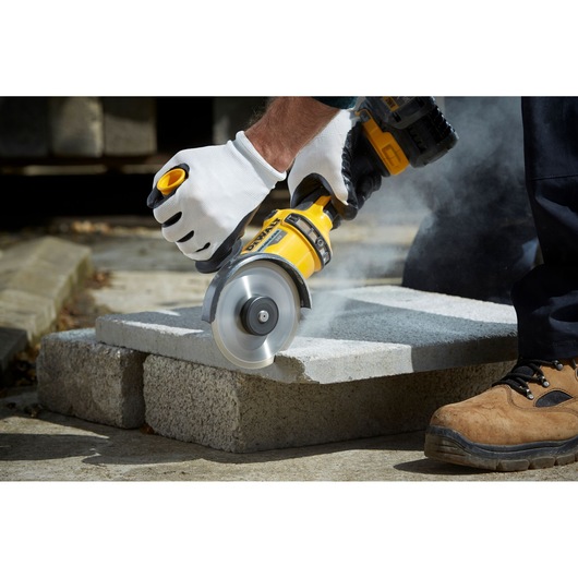 Person uses a DEWALT angle grinder to cut concrete blocks, creating dust. Safety gloves and boots are worn.