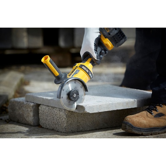 Person uses a DEWALT cutting tool to cut a concrete block outside. Tool is yellow and handheld.