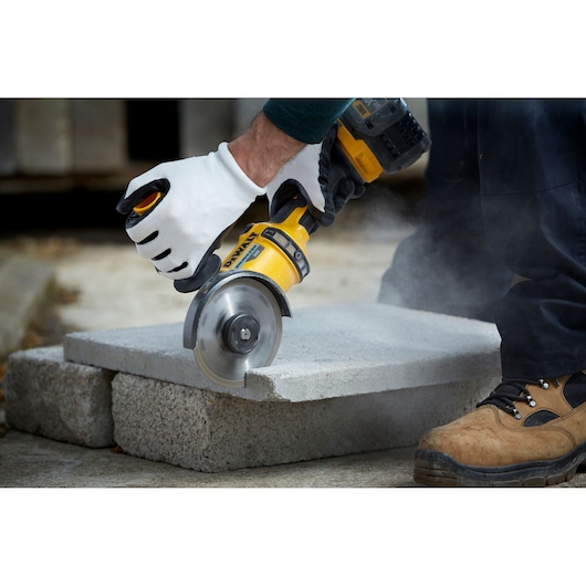A person wearing white work gloves uses a yellow DEWALT power tool to cut through a concrete slab, creating dust. The person