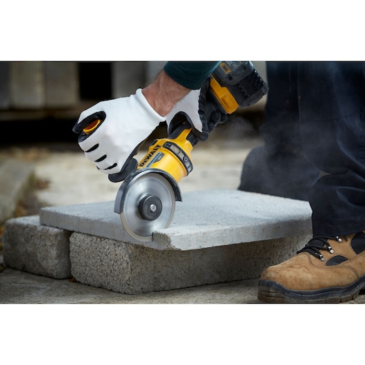 A person wearing white safety gloves uses a DEWALT power tool to cut a concrete slab outdoors. The tool is producing dust as