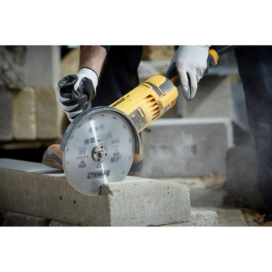 A person wearing gloves uses a yellow DEWALT angle grinder with a DT3732 diamond cutting disc to cut a concrete block. The